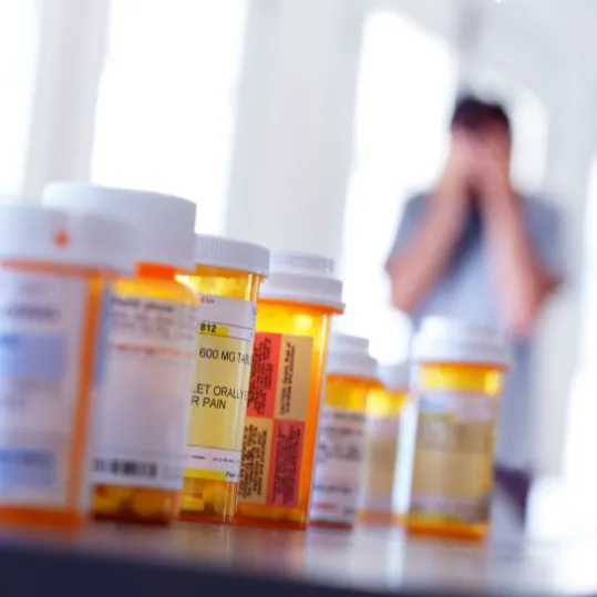 Prescription pill bottles in foreground with a distressed person in the background, representing medication-related mental health harm.